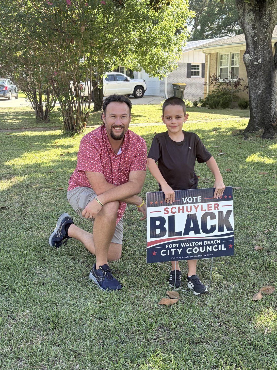 Schuyler Black kneeling in a Fort Walton Beach front yard beside a supporter's child, next to a campaign yard sign.